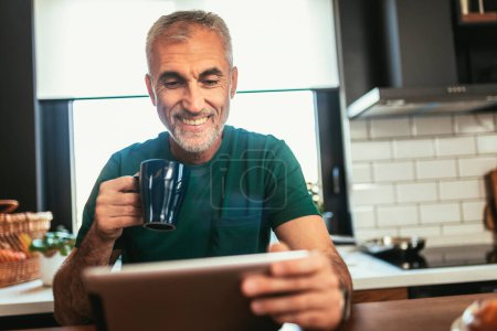 Téléchargez les photos : Homme mûr dans la cuisine travaillant à partir de la maison en utilisant une tablette numérique - en image libre de droit