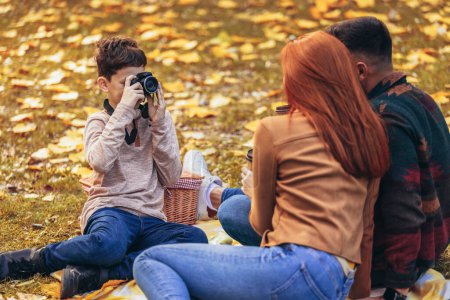 Foto de Jóvenes padres felices divirtiéndose con su hijo en el parque durante el día de otoño. Niño toma una foto de sus padres en un picnic - Imagen libre de derechos