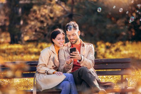 Téléchargez les photos : Jeune couple regardant leur tablette. Ils sont assis sur un banc lors d'une journée ensoleillée d'automne dans le parc coloré - en image libre de droit