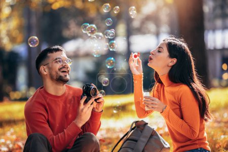 Foto de Novio tomando una foto de su novia mientras ella sopla burbujas. Día soleado de otoño en el colorido par - Imagen libre de derechos
