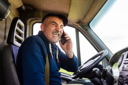 Téléchargez les photos : Mature camion conducteur à l'aide de téléphone mobile tout en conduisant véhicule de transport. - en image libre de droit