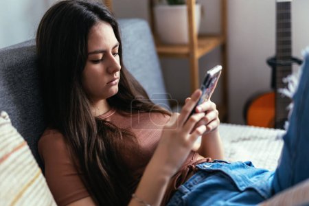 Téléchargez les photos : Beautiful teenager girl surfing social media on smart phone while having leisure time at her home. - en image libre de droit