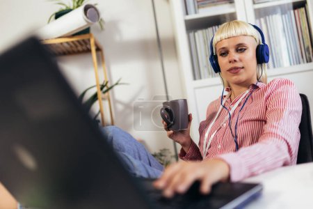 Téléchargez les photos : Jeune femme blonde travaillant sur son ordinateur portable et buvant du café - en image libre de droit