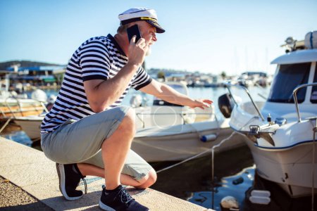 Foto de Hombre maduro parado cerca del mar vestido con una camisa y un sombrero de marinero usando un teléfono inteligente - Imagen libre de derechos