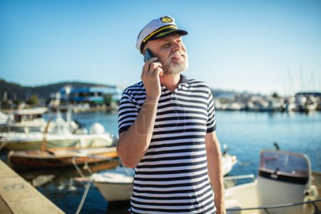 Téléchargez les photos : Homme mûr debout près de la mer vêtu d'une chemise et d'un chapeau de marin en utilisant un téléphone intelligent - en image libre de droit