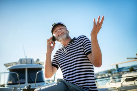 Foto de Hombre maduro parado cerca del mar vestido con una camisa y un sombrero de marinero usando un teléfono inteligente - Imagen libre de derechos