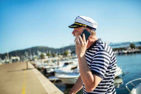 Photo for Mature man standing near the sea dressed in a sailor's shirt and hat using smart phone - Royalty Free Image