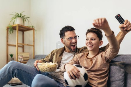 Photo for Father and son watching a soccer game while eating popcorn. - Royalty Free Image