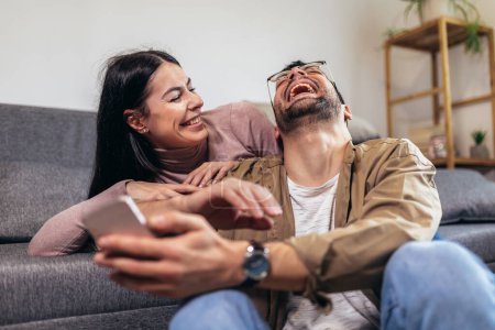 Téléchargez les photos : Beau jeune couple aimant coller les uns aux autres et faire du selfie tout en étant assis sur le canapé ensemble - en image libre de droit