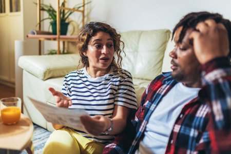 Téléchargez les photos : Couple multiracial à la maison analysant leurs finances avec des documents et un ordinateur portable. - en image libre de droit