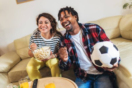 Foto de Esposo afroamericano y esposa latina viendo un partido de fútbol en la televisión desde la comodidad de su hogar. - Imagen libre de derechos