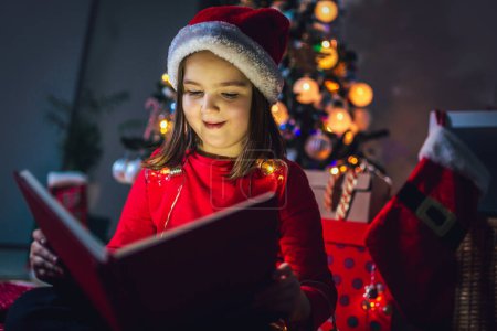 Photo for Little girl is reading a book on christmas. Colorful lights, Santa Claus hat and christmas tree - Royalty Free Image