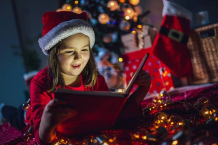 Foto de La niña está leyendo un libro sobre Navidad. Luces coloridas, sombrero de Papá Noel y árbol de Navidad - Imagen libre de derechos