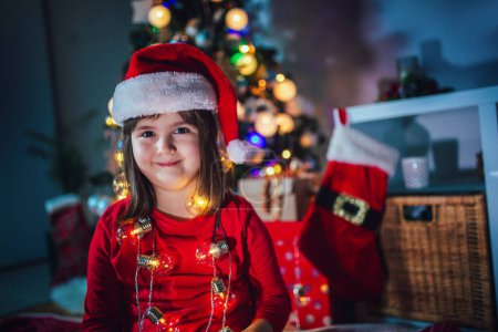 Foto de La niña está sonriendo en la cámara en Navidad. Luces coloridas, sombrero de Papá Noel y árbol de Navidad - Imagen libre de derechos