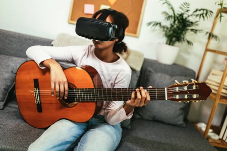 Foto de African american teenage girl sitting on couch in her room and learning to play guitar using VR glasses to immerse herself in metaverse - Imagen libre de derechos