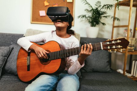 Téléchargez les photos : African american teenage girl sitting on couch in her room and learning to play guitar using VR glasses to immerse herself in metaverse - en image libre de droit