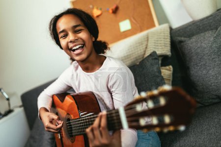 Foto de African american teenage girl sitting on couch in her room and learning to play guitar - Imagen libre de derechos