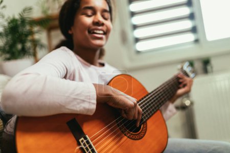 Photo for African american teenage girl sitting on couch in her room and learning to play guitar - Royalty Free Image