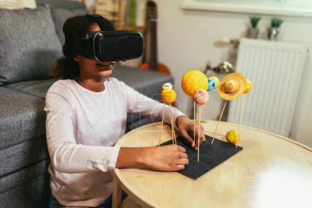Photo for Happy african american school girl making a solar system for a school science project at home using VR glasses to immerse herself in metaverse - Royalty Free Image
