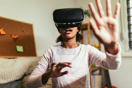 Téléchargez les photos : African American girl using virtual reality glasses in her room. Black girl enjoys VR technology to watch 3D movies or immerse herself in metaverse - en image libre de droit