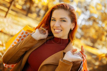 Téléchargez les photos : Ginger girl revenant du shopping avec des sacs. Parcourir un parc coloré par une journée d'automne ensoleillée. - en image libre de droit