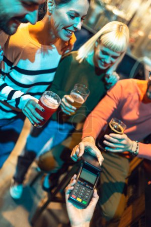 Téléchargez les photos : Diverse friends in bar paying with cellular device - en image libre de droit