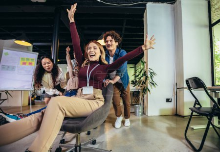 Téléchargez les photos : Jeunes hommes d'affaires joyeux en tenue décontractée intelligente s'amusant en course sur des chaises de bureau et souriant - en image libre de droit