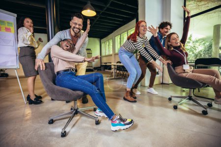 Photo for Young cheerful businesspeople in smart casual wear having fun while racing on office chairs and smiling - Royalty Free Image