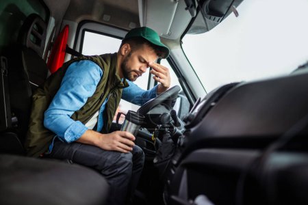 Téléchargez les photos : Chauffeur de camion fatigué tenant du café dans sa cabine. - en image libre de droit