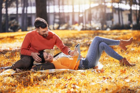 Téléchargez les photos : Jeune homme et femme assis sur une couverture dans le parc à l'automne à l'aide d'une tablette numérique. - en image libre de droit