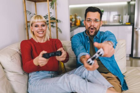 Téléchargez les photos : Petit ami et petite amie jouer à un jeu vidéo avec des joysticks dans le salon. Couple aimant jouent à des jeux vidéo à la maison - en image libre de droit