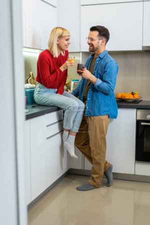 Foto de Joven hombre y mujer de pie en la cocina y hablando. Feliz esposa y marido con tazas de café y jugo de pie en la cocina. - Imagen libre de derechos