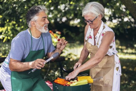 Photo for Elderly couple are making barbeque, vegetables, and drinking beverages while enjoying and making memories. - Royalty Free Image
