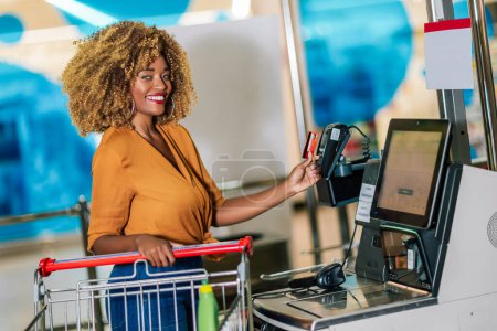 Foto de African American Woman with bank card buying food at grocery store or supermarket self-checkout - Imagen libre de derechos