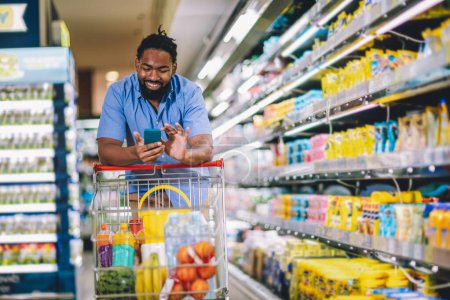 Téléchargez les photos : Black Male Buyer Shopping Groceries In Supermarket Taking Product From Shelf Standing With Shop Cart Indoors Using Phone. - en image libre de droit