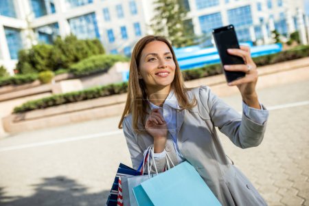 Téléchargez les photos : Attrayant jeune femme prenant des photos sur son téléphone portable à l'extérieur tout en faisant du shopping dans la ville - en image libre de droit