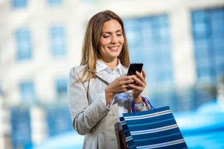 Foto de Mujer joven atractiva usando su teléfono celular afuera mientras hace compras en la ciudad - Imagen libre de derechos