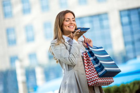 Foto de Atractiva joven mujer que envía mensajes de voz en su teléfono celular mientras hace compras en la ciudad - Imagen libre de derechos