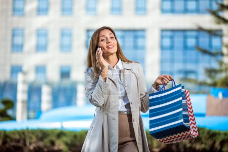 Foto de Atractiva joven mujer hablando en su teléfono celular afuera mientras hace compras en la ciudad - Imagen libre de derechos