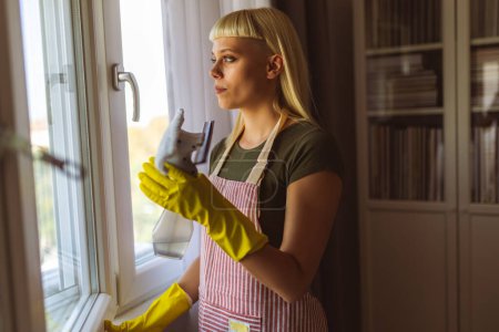 Téléchargez les photos : Jeune femme blonde nettoyer les fenêtres dans sa maison - en image libre de droit