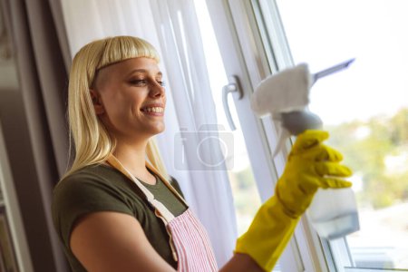 Téléchargez les photos : Jeune femme blonde nettoyer les fenêtres dans sa maison - en image libre de droit