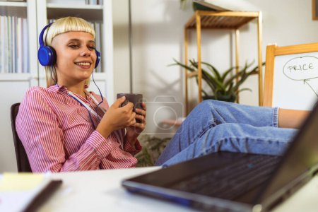 Téléchargez les photos : Jeune femme blonde écoutant de la musique sur son ordinateur portable et buvant du café - en image libre de droit