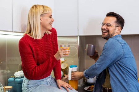 Photo for Young man and woman standing in kitchen and talking. Happy wife and husband with cups of coffee and juice standing in the kitchen. - Royalty Free Image