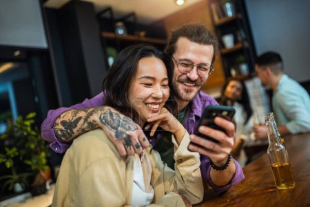 Photo for Two diverse young friends laughing together and looking at the phone during a dinner together in a friend's kitchen - Royalty Free Image