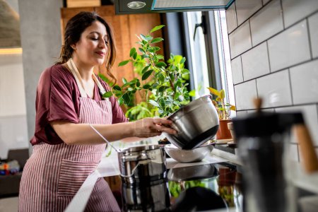 Téléchargez les photos : Latino femme cuisine dans sa cuisine moderne en remuant dans le pot faire de la nourriture pour le dîner. - en image libre de droit