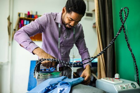 Téléchargez les photos : Jeune homme afro-américain tailleur chemise de repassage dans son atelier. - en image libre de droit