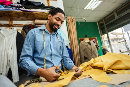 Photo for Young African American tailor cuts with scissors yellow fabric in his own atelier. - Royalty Free Image