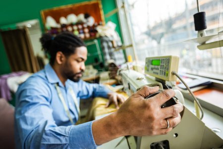 Téléchargez les photos : Jeune tailleur afro-américain mâle coudre avec une machine à coudre dans son propre atelier - en image libre de droit