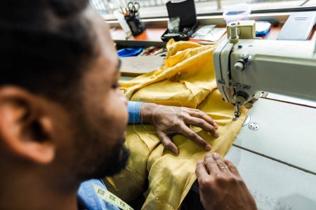 Téléchargez les photos : Jeune tailleur afro-américain mâle coudre avec une machine à coudre dans son propre atelier - en image libre de droit