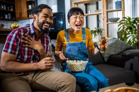 Foto de Pareja afroamericana y asiática, en el sofá viendo televisión, bebiendo cerveza y comiendo palomitas de maíz y pizza. - Imagen libre de derechos